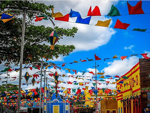 Foto da praça de uma típica cidade pequena do interior com casas coloridas que variam entre tons de azul e tons de amarelo e nesta praça possuem postes na cor cinza parte de uma arvore ao lado esquerdo, um céu azul com nuvens e bandeirinhas e balões em tons de amarelo, azul verde e vermelho. colocados sobre toda a praça e cruzando a rua.