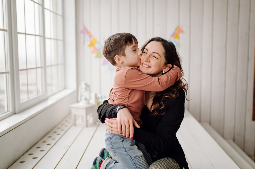 Foto de um filho de blusa vermelha, calça jeans e meia colorida, abraçando e beijando sua mãe de blusa preta e calça quadriculada. Dentro de um quarto infantil de cor branca, com uma grande janela na cor branca ao lado ao fundo alguns brinquedos e na parede ao fundo alguma bandeirinhas coloridas.